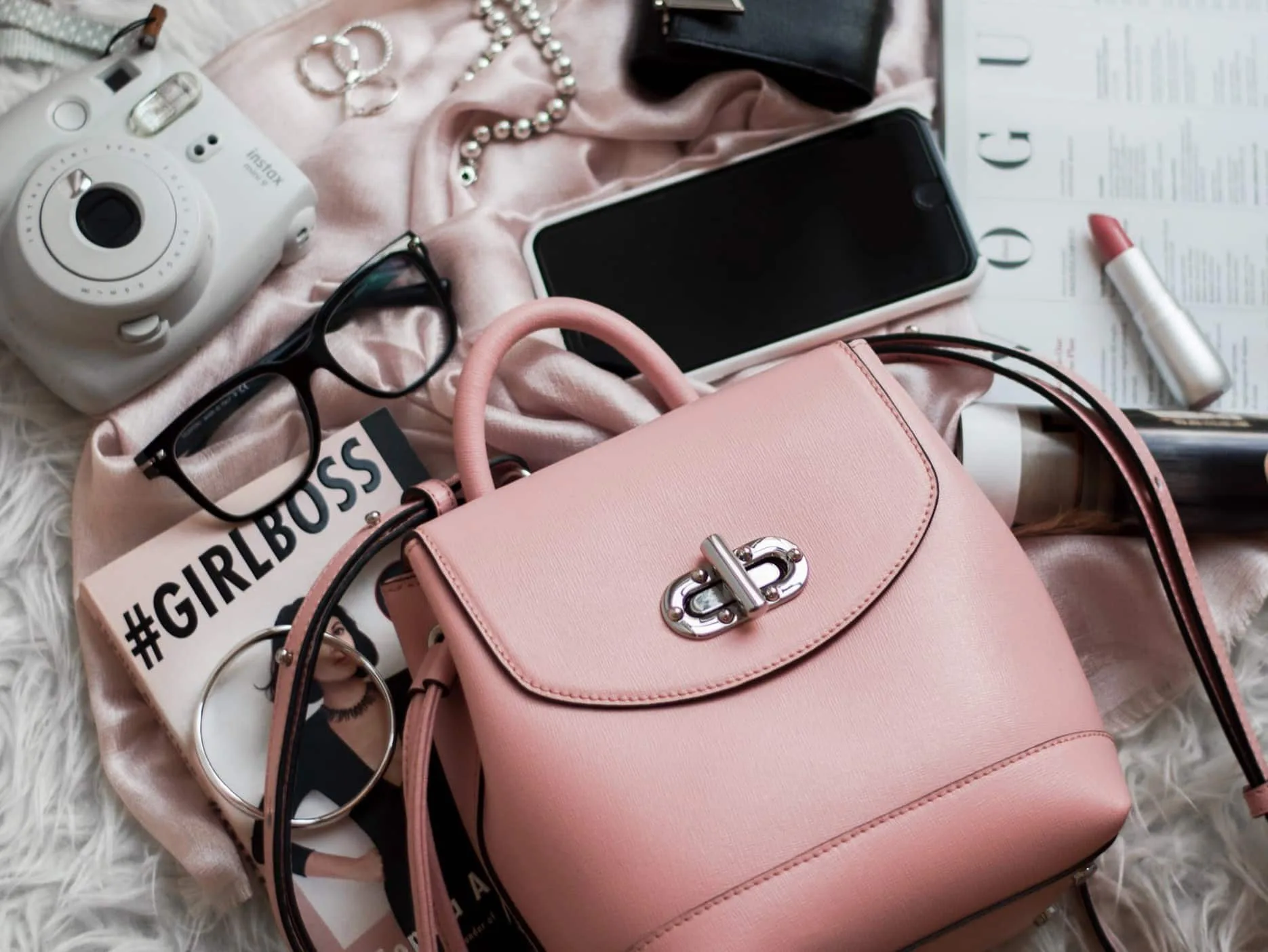 Różowy plecak, okulary, telefon, aparat Instax, biżuteria, pomadka i magazyn, rozłożone na miękkiej powierzchni.