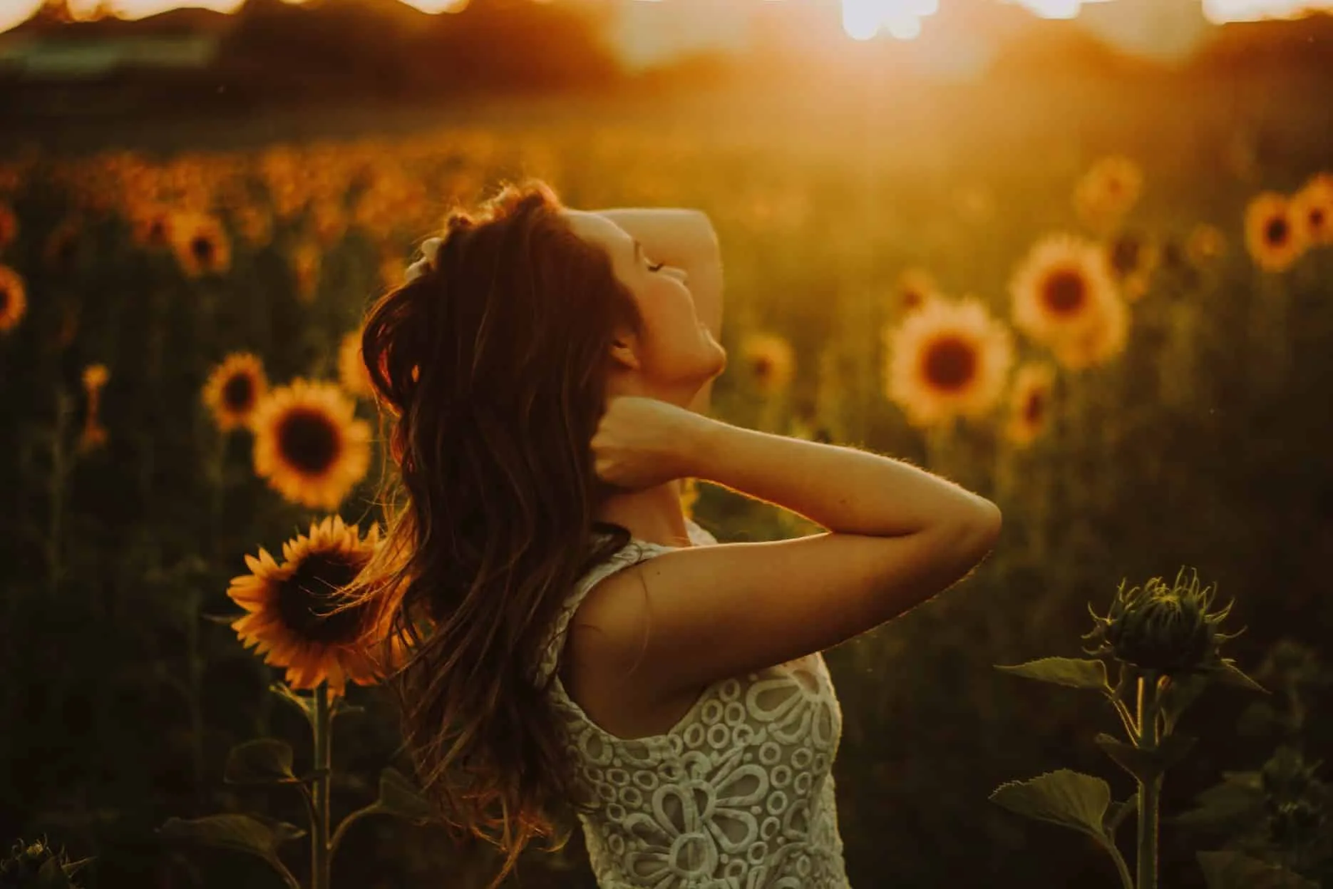 Kobieta stojąca wśród pola słoneczników, ciesząca się zachodem słońca, z ręką za głową w promieniach wieczornego światła.