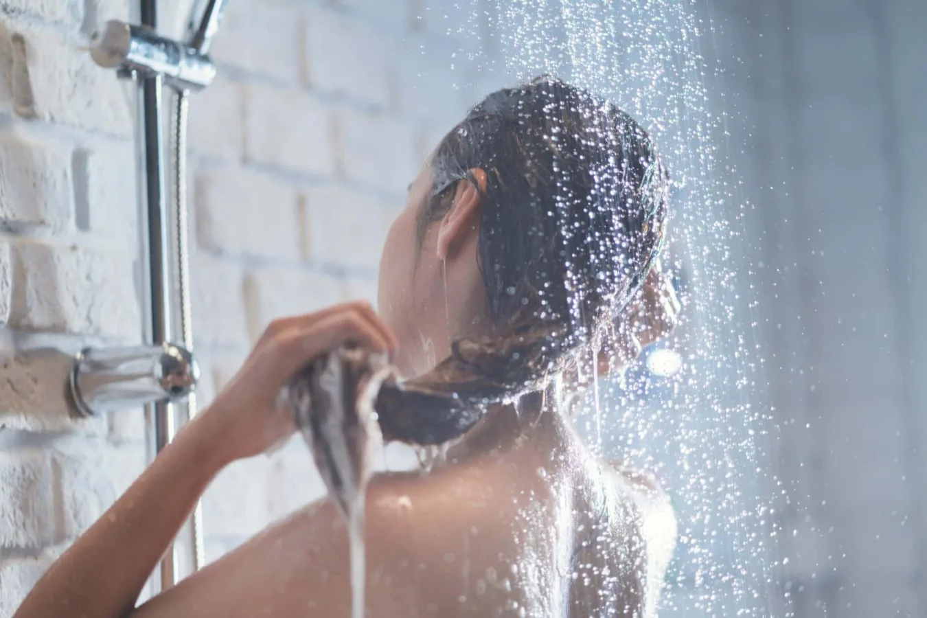 Kobieta myjąca włosy pod prysznicem, z kroplami wody rozpryskującymi się wokół.
