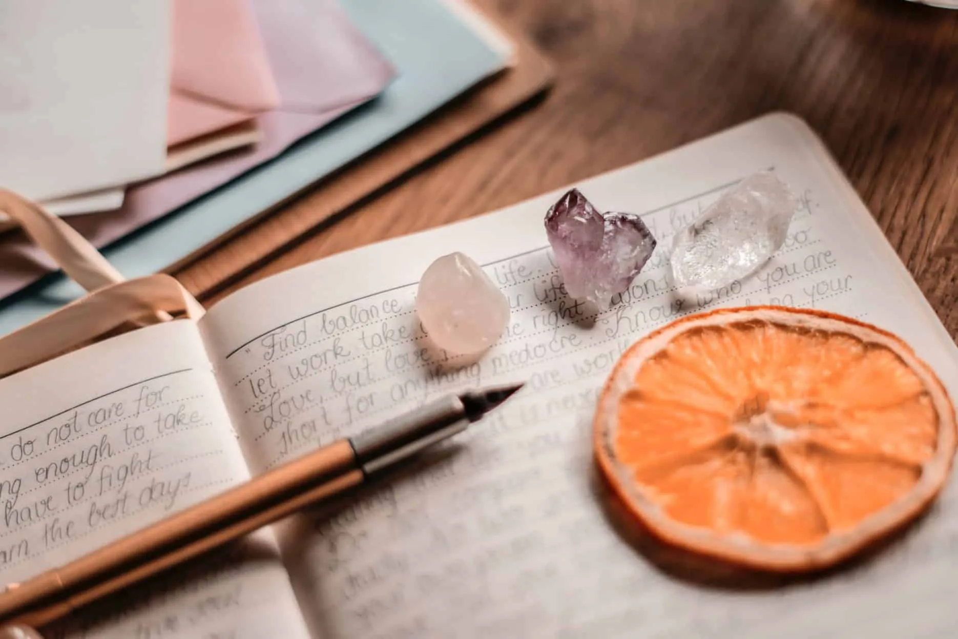Notatnik z ręcznie pisanych refleksji, obok pióra, kryształów i suszonego plasterka pomarańczy na drewnianym stole.
