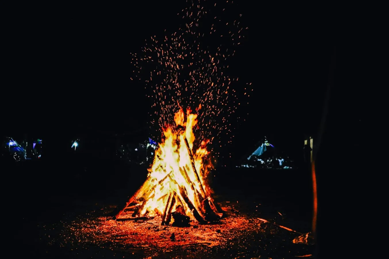 Ognisko z iskrami w nocy, z sylwetką namiotu w tle.